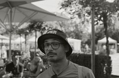 Black and white image of a man in a bucket hat and large glasses, his head slightly tilted up with a skeptical look on his face. He appears casual, with sunglasses hanging on the shirt around his neck.