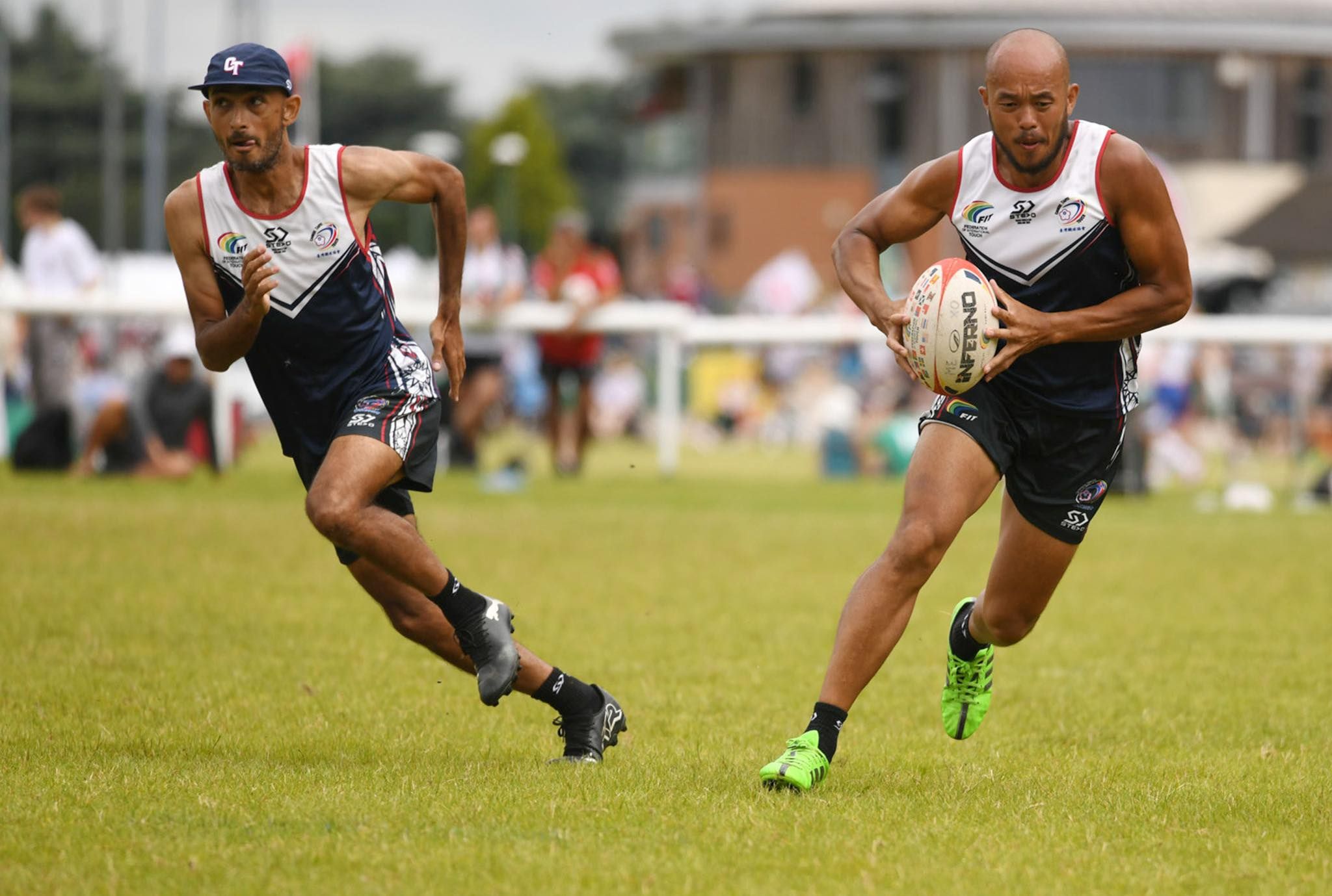Two men running towards the camera angling and looking in opposite directions. They are both in sports uniforms, with sleeveless shirts. One is holding a rugby ball, while the other runs besides him in support.